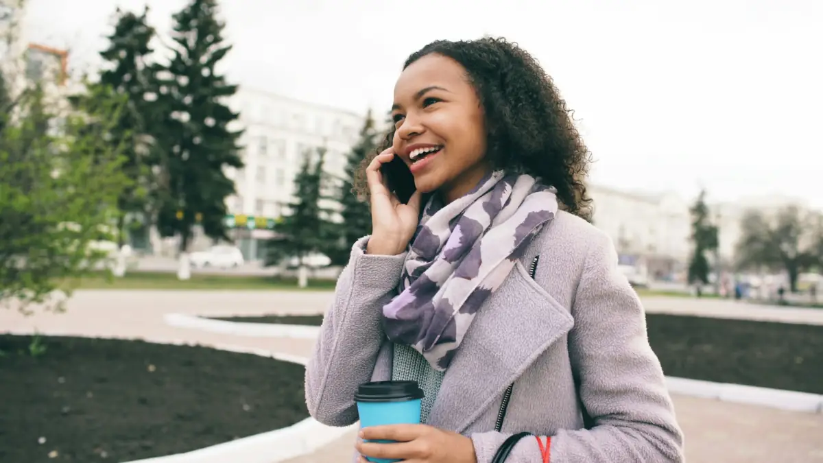 A smiling woman talking on her phone outdoors, holding a takeaway coffee.