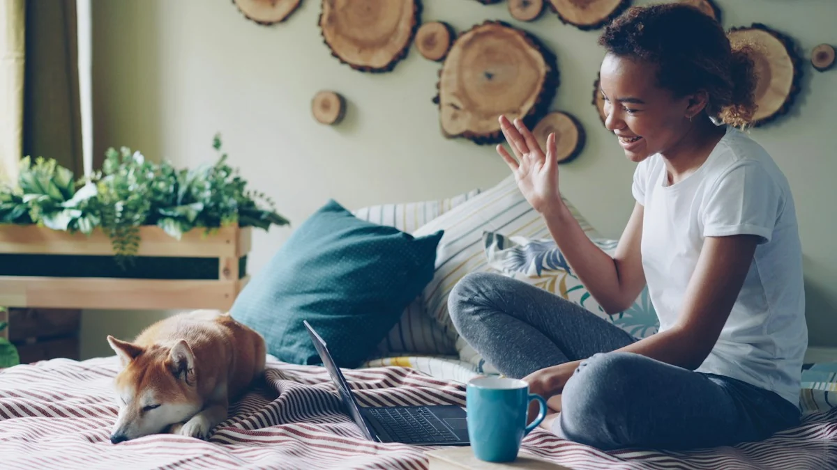 A woman at home waving at her laptop during a call, with her dog resting beside her.