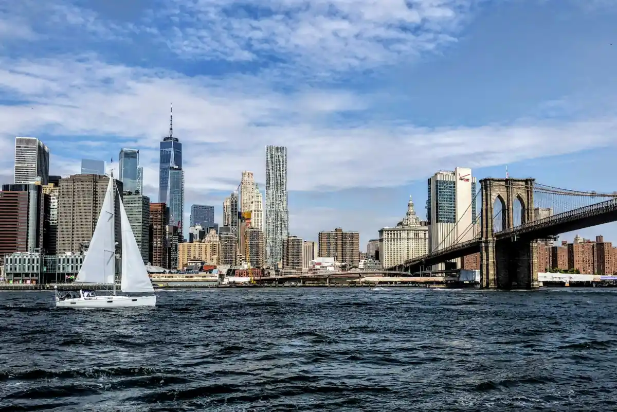 A sailboat on the East River in front of the Lower Manhattan skyline and the Brooklyn Bridge on a sunny day.