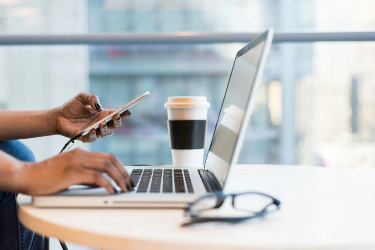 Someone at a cafe table checking a smartphone while working on a laptop, a coffee cup beside them.